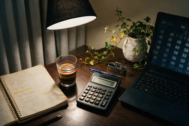 Mesa de trabalho à noite com caderno de contas feitas à mão, calculadora e um notebook ao lado